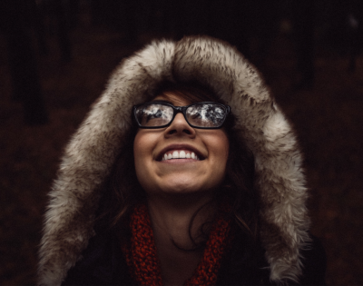A woman wearing a fur hood and glasses looks up toward an unseen object outside of the camera's view. She is smiling.