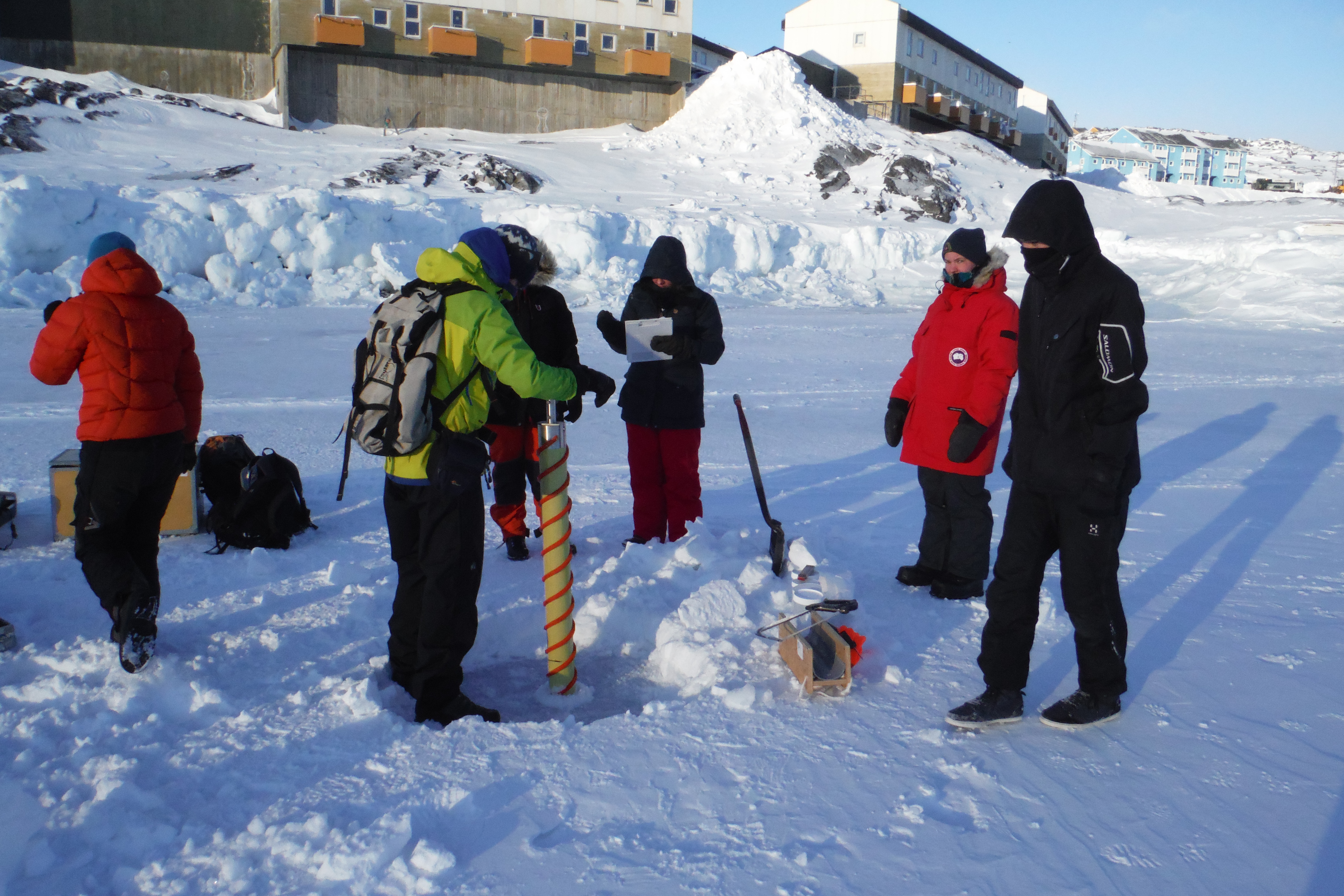 Students extract an ice core from Nuuk Old Harbour using the Kovacs corer (the long orange barrel, centre). This corer is being hand-turned, but they can be driven by power tools for greater speed, and can be used with extensions to core much thicker ice. Photo credit: Maxim Lamare