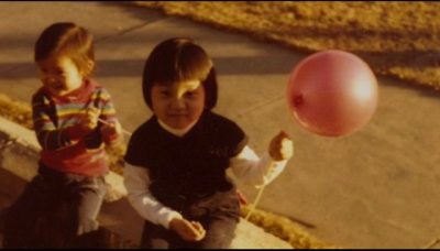 still shot from film of two children sitting on the sidewalk, looking at the camera