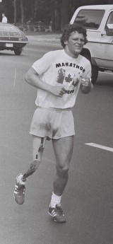 Terry Fox, a young man with short, curly hair and an artificial right leg runs down a street. He wears shorts and a T-shirt that reads "Marathon of Hope"