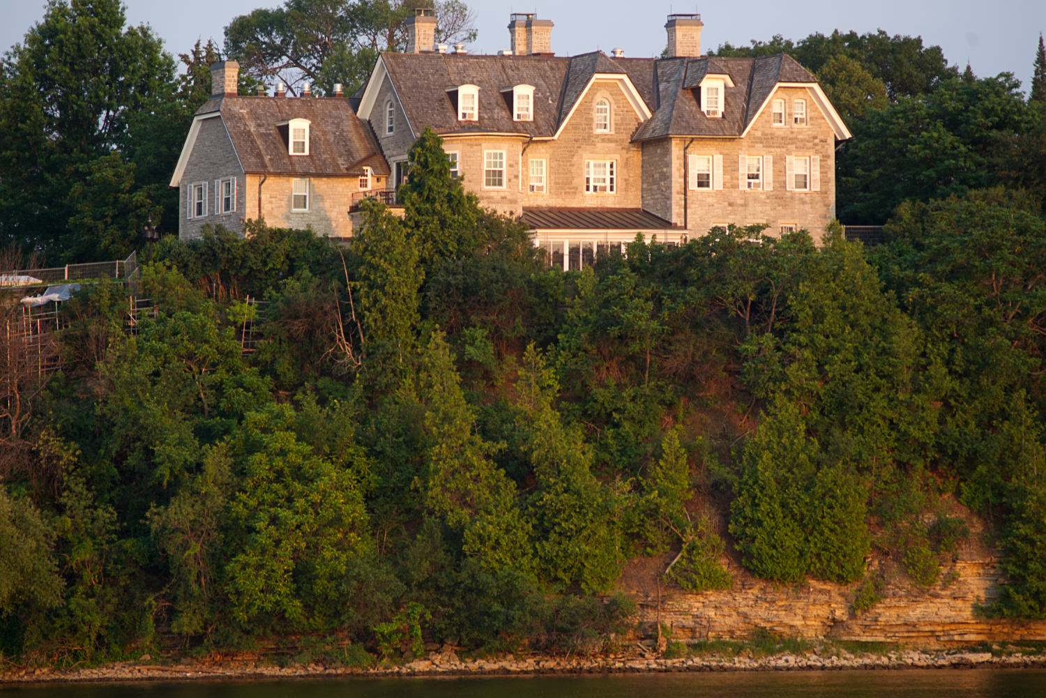 A sunlit limesone building sits atop a cliff above a river.
