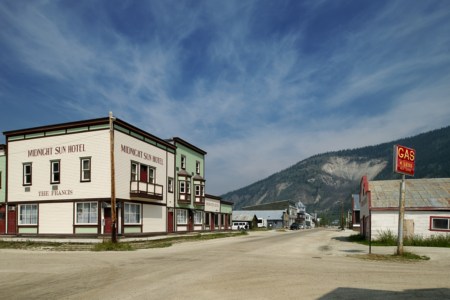 A streetscape of a wide, dirt street, with antiquated wooden buildings on either side and mountains in the background.