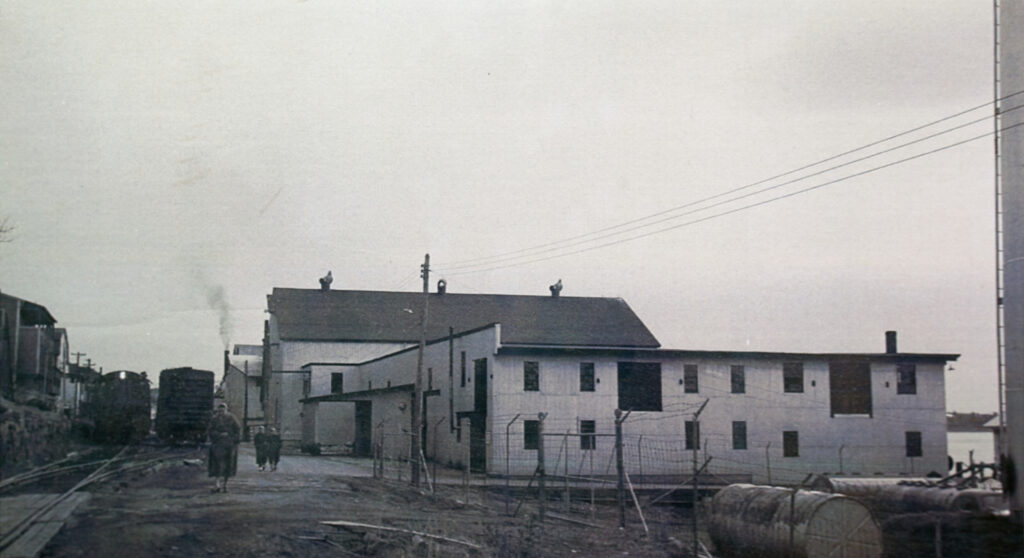 An archival B&W photo showing an ensemble of broad wooden buildings with train tracks running along one side.