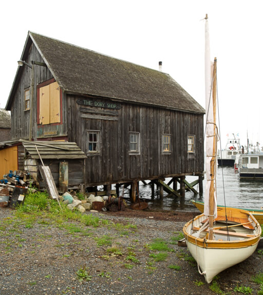 A small wooden shed propped above the water on stilts, with a sign reading “The Dory Shop”.