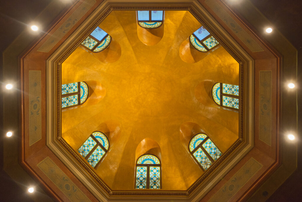 View straight up into a lofty dome with an octagonal base and eight stained glass windows.
