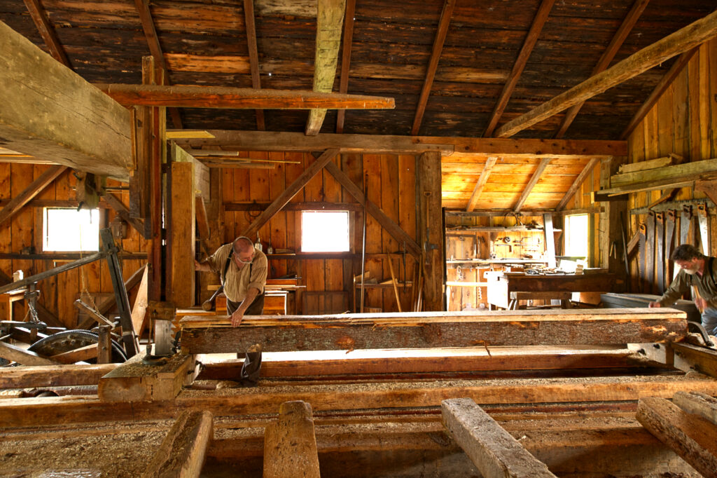 Two men work with a very bog log inside a wooden shed. The log is laid flat and metal machinery is clustered at one end.