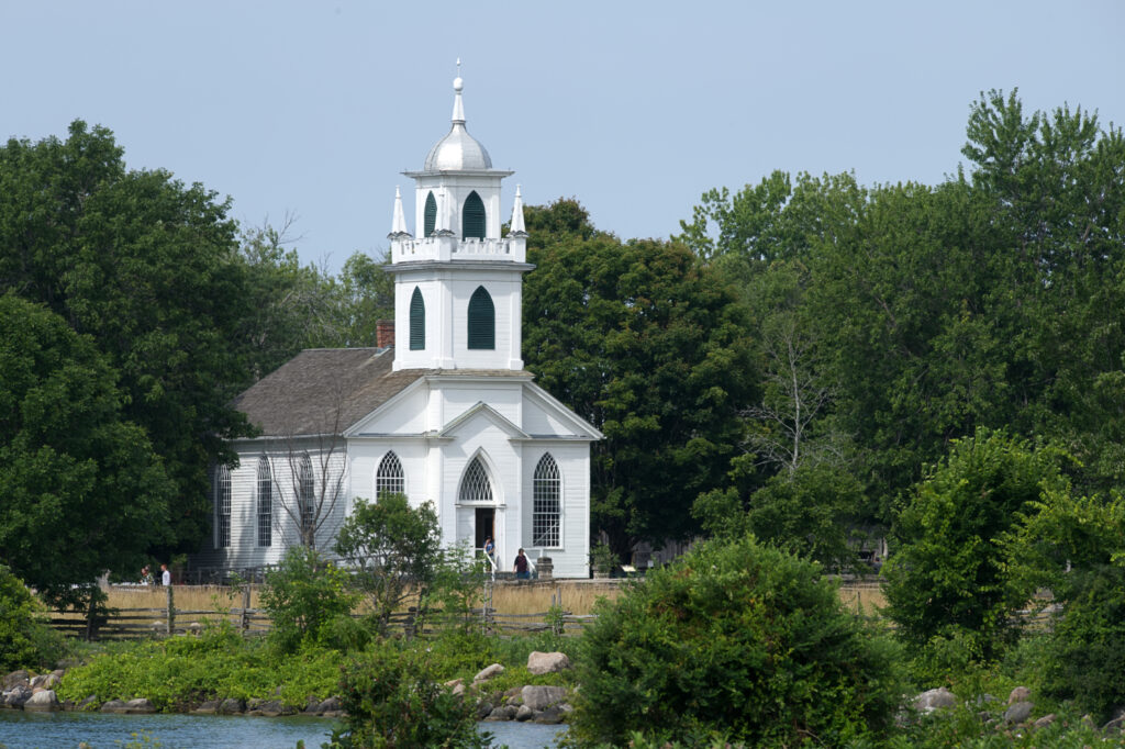 A white wooden church with a decorative front tower sits in a picturesque landscape of trees, rocks, and water.