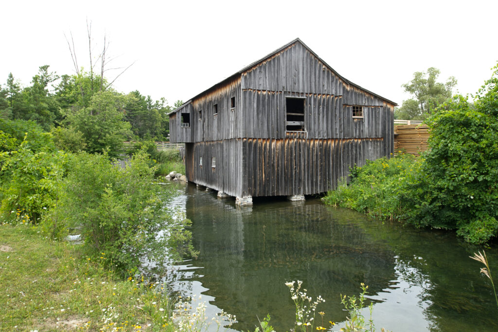 A large but very simple and rustuc wooden shed sits above a small body of water.