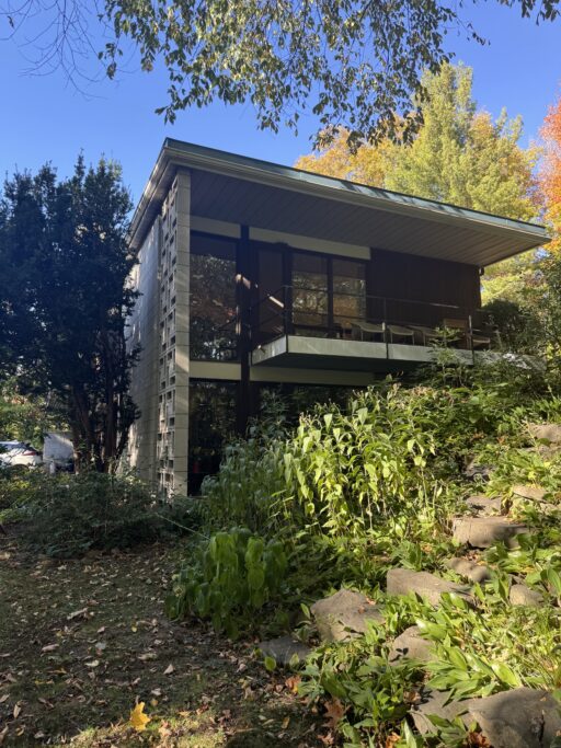 An angular Modernist house with a deeply projecting balcony and eves sits on a plant-filled hill.