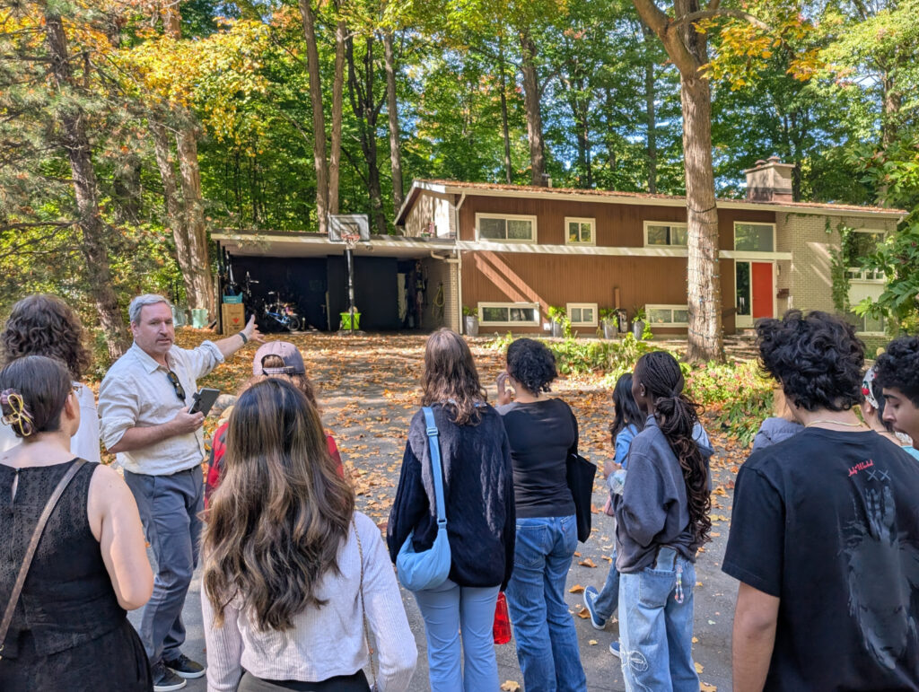 Tour leader Andrew Waldron speaks to students as he gestures toward aa wood-finished Modern house.