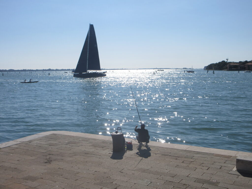 A lone seated fisherman casts into the water with a sailboat an some small vessels beyond.