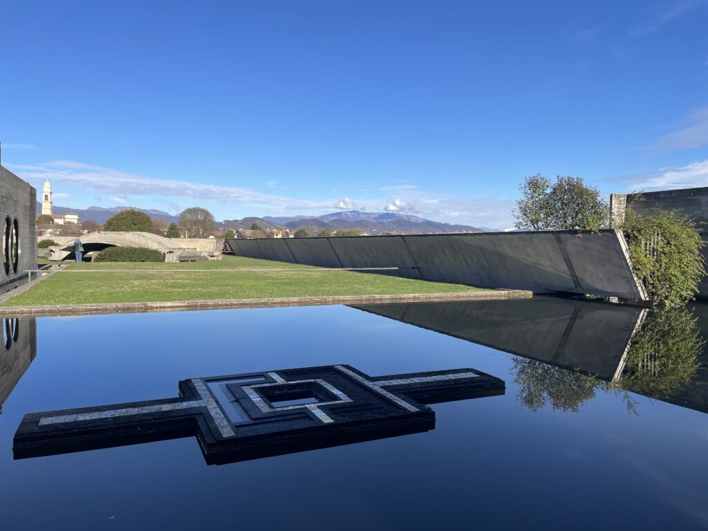 Sky and sculpted concrete forms are reflected in the mirror-like surface of a pool of water.