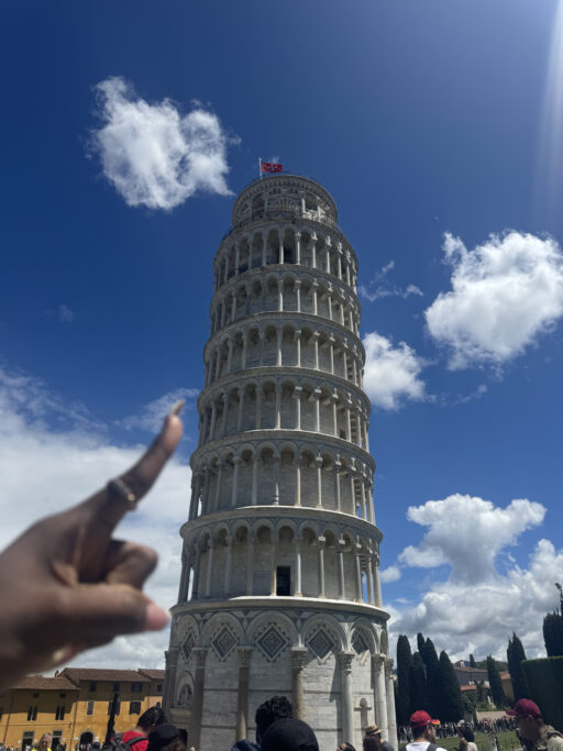 A human hand at the left edge of the frame points to the Leaning Tower of Pisa.