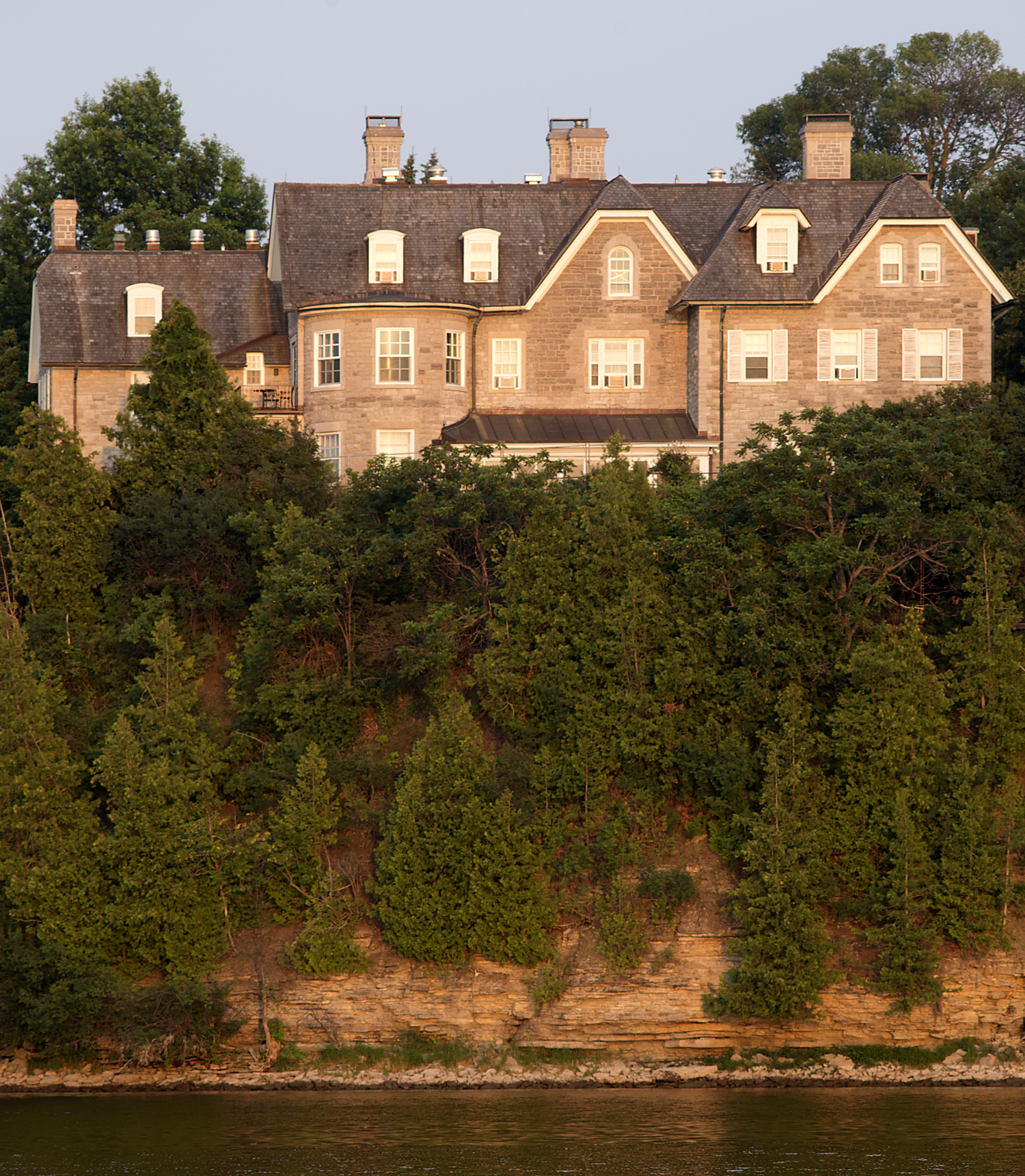 Golden sunlight illuminates a limestone building sitting atop an escarpment over a river.