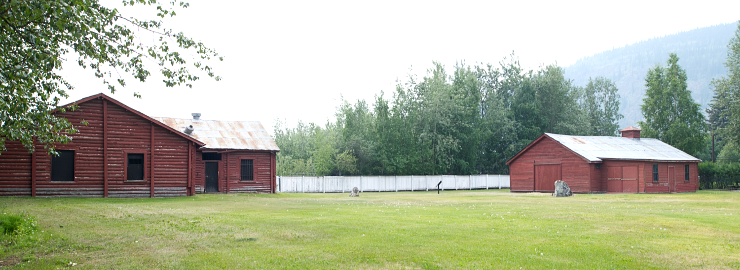 Two fairly large log buildings sit in an open, grassy area.