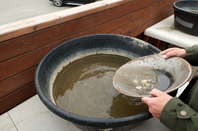 A collection of small stones and sand is swished around in a large, hand-held metal pan.