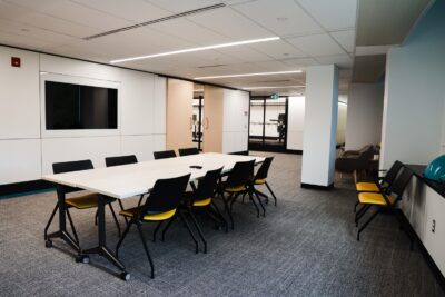 Abilities Living Laboratory common area. A large open space, showing a large table with eight chairs around it. There is a large screen embedded in the far wall.