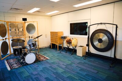 We Are All Musicians Laboratory showing various musical instruments including a drum set, a large gong, and a wall of gongs with electronics solenoids used to hit them.