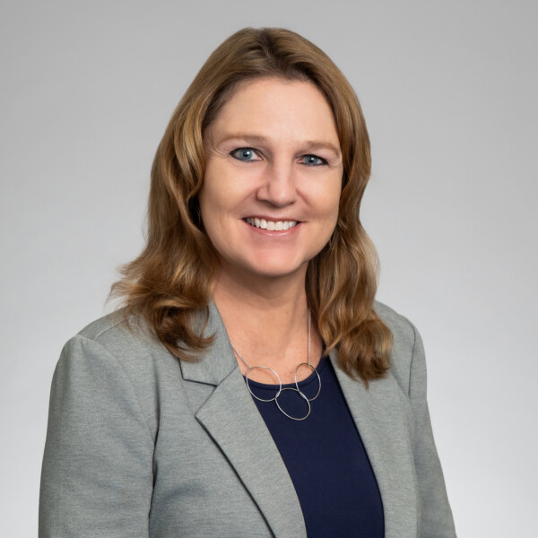 Portrait of a smiling woman with shoulder-length brown hair, wearing a grey blazer, navy top and silver necklace, against a light grey background.