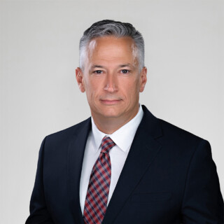A professional headshot of a man wearing a dark suit, and a dark red tie.