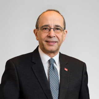 A professional headshot of a man wearing glasses, with a dark suit, a grey and blue tie, and a red pin on the lapel of his suit jacket.