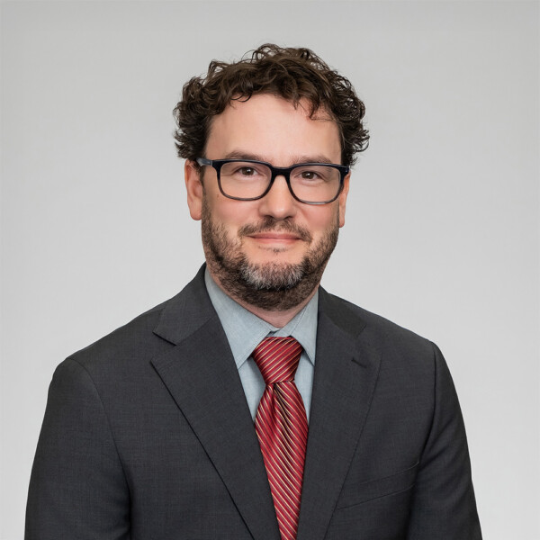 A professional headshot of a man with glasses, wearing a dark suit with a red tie.