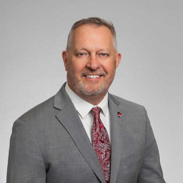 A professional headshot of a man wearing a grey suit, with a pin on the lapel of the suit.