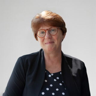 A photo of a woman with glasses and short red hair, wearing a dark jacket and a dark shirt with white polka dots.