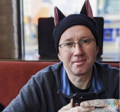 A white, middle-aged man wearing glasses and a black hat with cat ears sits at a table in a cafe, holding a smartphone.