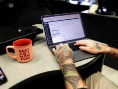 A white man with tattooed arms typing on a laptop at a desk, with a mug labeled “Best Cat Mom Ever” nearby.