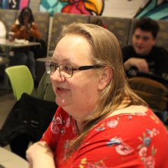 A middle-aged white woman with blonde hair and glasses sits in a coffee shop, chatting.