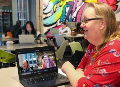 A middle-aged white woman with blonde hair and glasses. She is sitting at a table with a laptop at Rooster's Coffeehouse. On the laptop screen are two smiling men in a Zoom meeting.