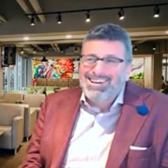 A middle-aged white man with glasses and a greying beard sits in a coffee shop, smiling.