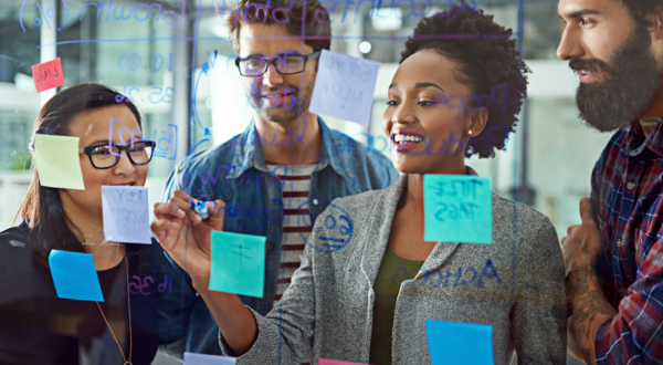 A group of people collaborate around a glass wall covered with handwritten notes and colorful sticky notes, discussing ideas during a brainstorming session in an office setting.