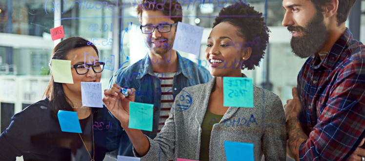 A group of people collaborate around a glass wall covered with handwritten notes and colorful sticky notes, discussing ideas during a brainstorming session in an office setting.