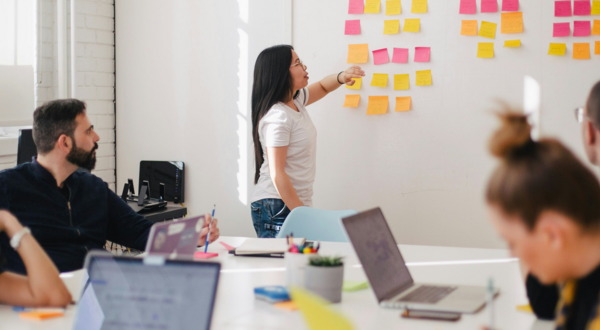 A person stands at a wall covered with colorful sticky notes, pointing during a collaborative meeting while others seated around a table listen and work on laptops in a bright office space.