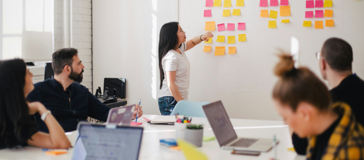 A person stands at a wall covered with colorful sticky notes, pointing during a collaborative meeting while others seated around a table listen and work on laptops in a bright office space.
