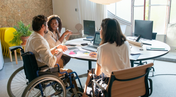 Three people participate in a collaborative meeting around a table in a bright office space, including one person using a wheelchair, with laptops, notes, and office materials visible throughout the room.
