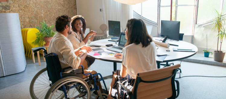 Three people participate in a collaborative meeting around a table in a bright office space, including one person using a wheelchair, with laptops, notes, and office materials visible throughout the room.