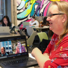 A middle-aged white woman with blonde hair and glasses. She is sitting at a table with a laptop at Rooster's Coffeehouse. On the laptop screen are two smiling men in a Zoom meeting.