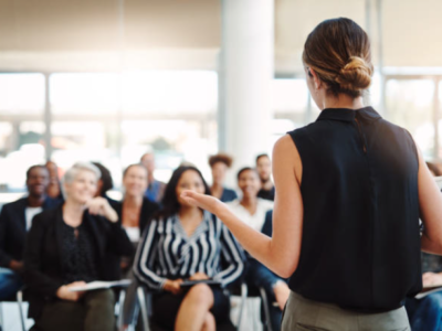 A woman stands with her back to us and faces a classroom of people in front of he as she delivers a talk or presentation to the crowd. Her brown and blonde hair is twisted into a neat, chic bun at the base of her head and she wears a coloured, sleeveless blouse and sage green trousers.