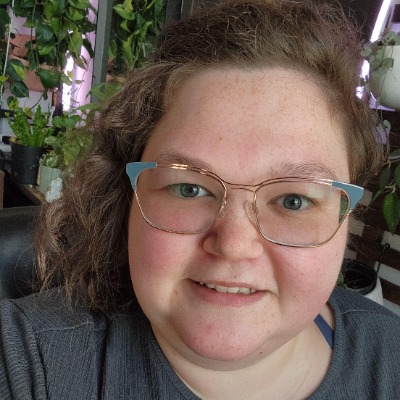 Woman wearing blue glasses and a grey shirt with short hair on one side and shoulder length hair on the other side.