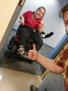 A Carleton student using a mobility chair poses behind the threshold of a doorway wearing a big smile. Half of Pascal Dubuc’s body appears inside the frame, indicating that they are taking a selfie together with the entrance threshold. Both the student and Pascal are giving a thumbs-up.
