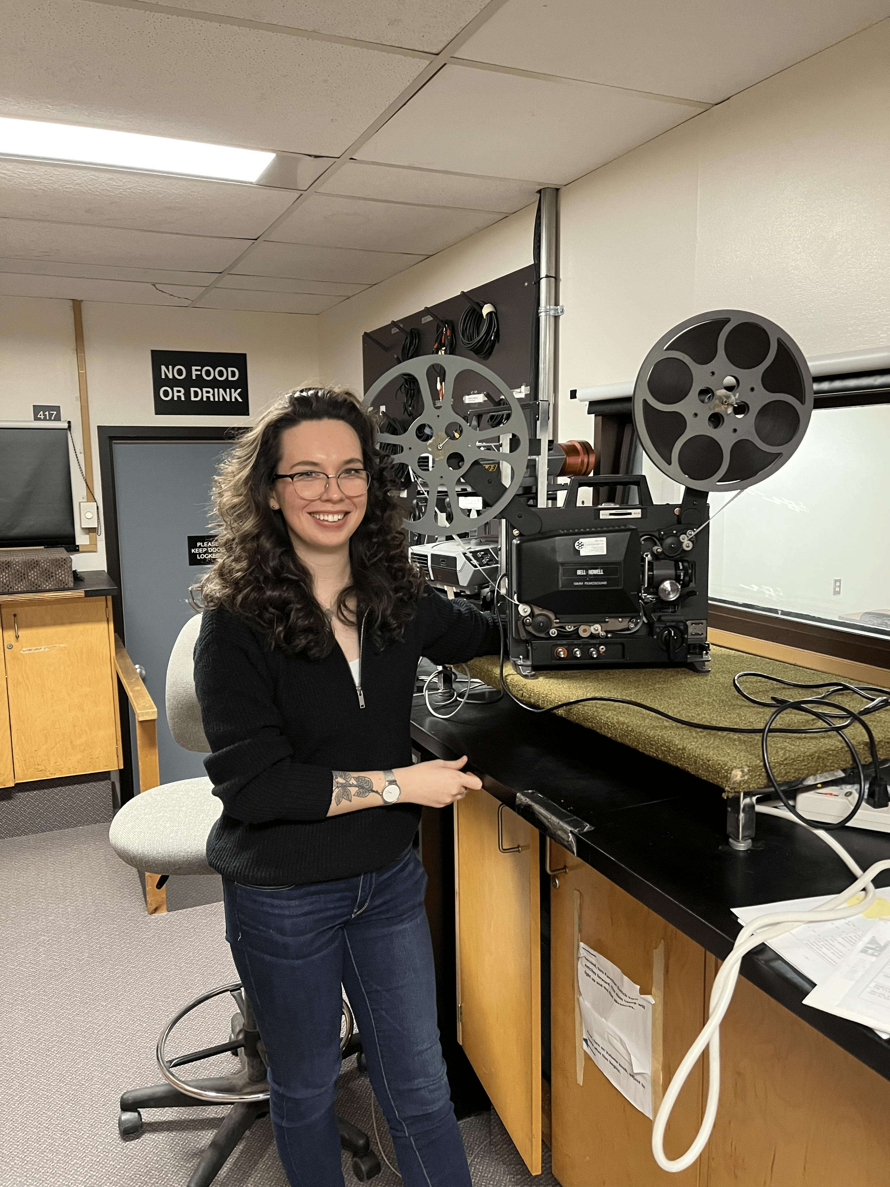 A photo of the author of the post, a young light-skinned person with long, curly, brown hair. They are posing with a piece of analogue film equipment.