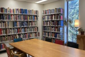 A corner of the main space of the AVRC with a window, bookshelves and a lamp along the walls, and a wooden table with chairs.