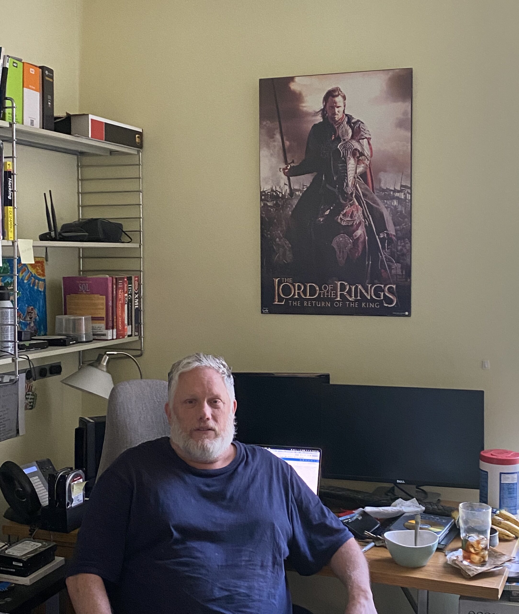 Photo of Adam, a middle-aged light-skinned man with graying hair. He is wearing a dark blue shirt and seated at his desk. On the wall there are shelves and a Lord of the Rings poster.