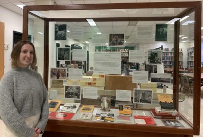 A photo of the author of the post, a young light-skinned person with long dirty blond hair, posing in front of a display case with material from the ACWA collection.