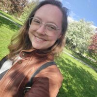 A photo of Elzi, a young light-skinned person with long dirty blonde hair and glasses. Elzi is smiling, wearing a brown leather jacket, and standing in front of a field and several trees in bloom.