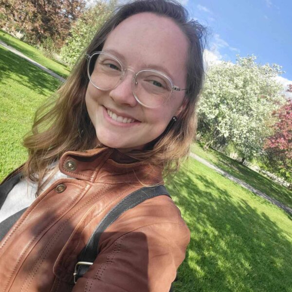 A photo of Elzi, a young light-skinned person with long dirty blonde hair and glasses. Elzi is smiling, wearing a brown leather jacket, and standing in front of a field and several trees in bloom.