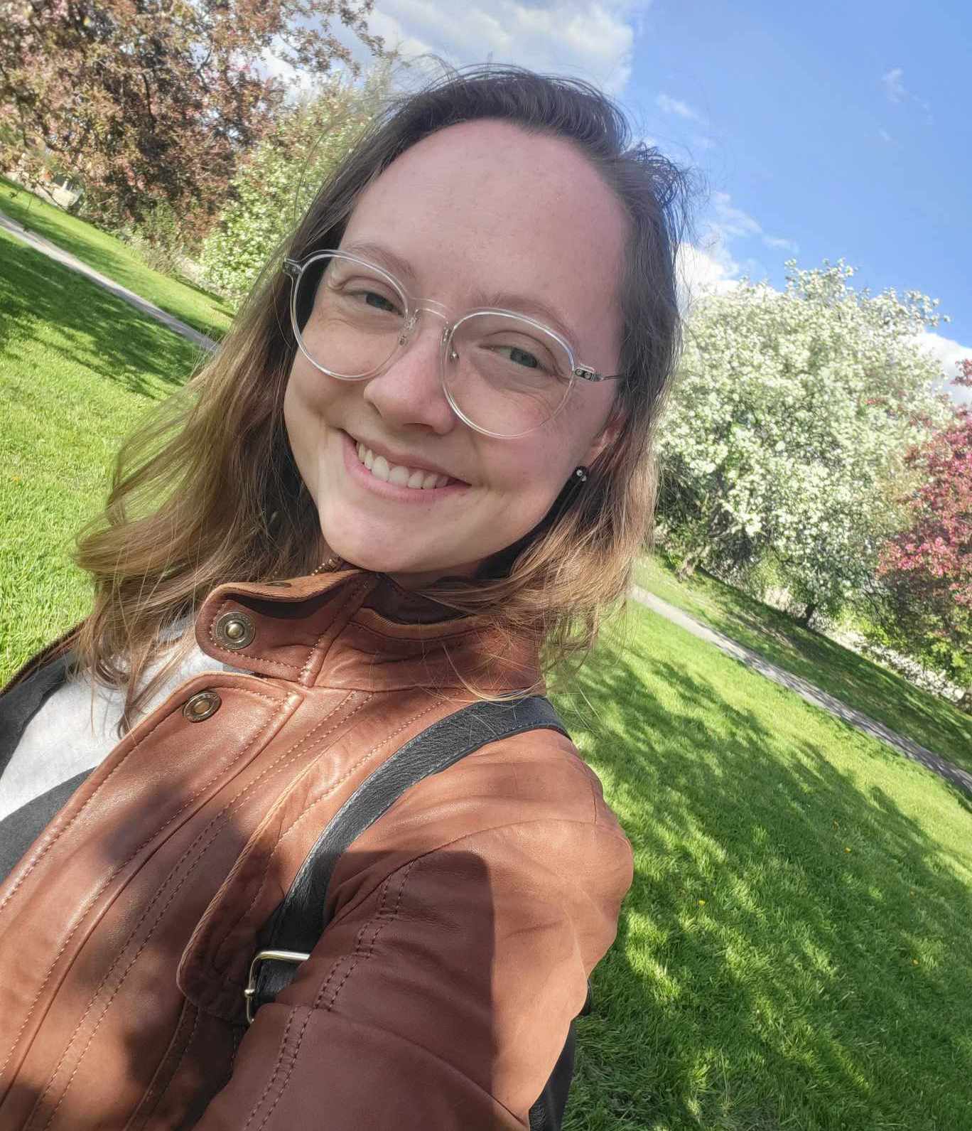 A photo of Elzi, a young light-skinned person with long dirty blonde hair and glasses. Elzi is smiling, wearing a brown leather jacket, and standing in front of a field and several trees in bloom.
